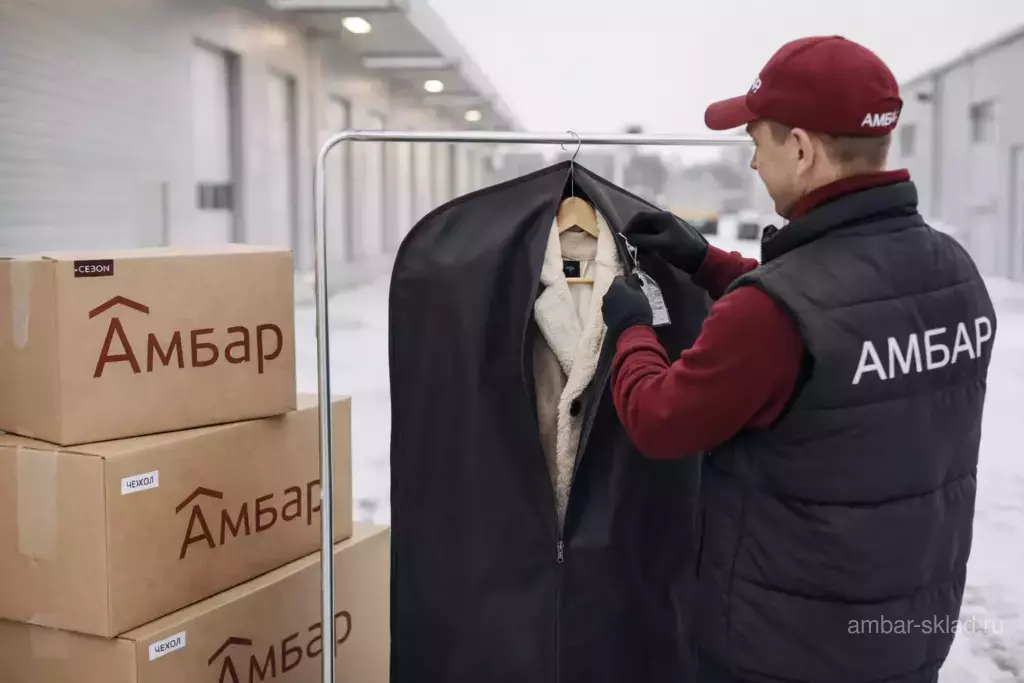 Сотрудник АМБАР на зимней уличной зоне склада убирает дубленку в тканевый чехол для правильного длительного хранения